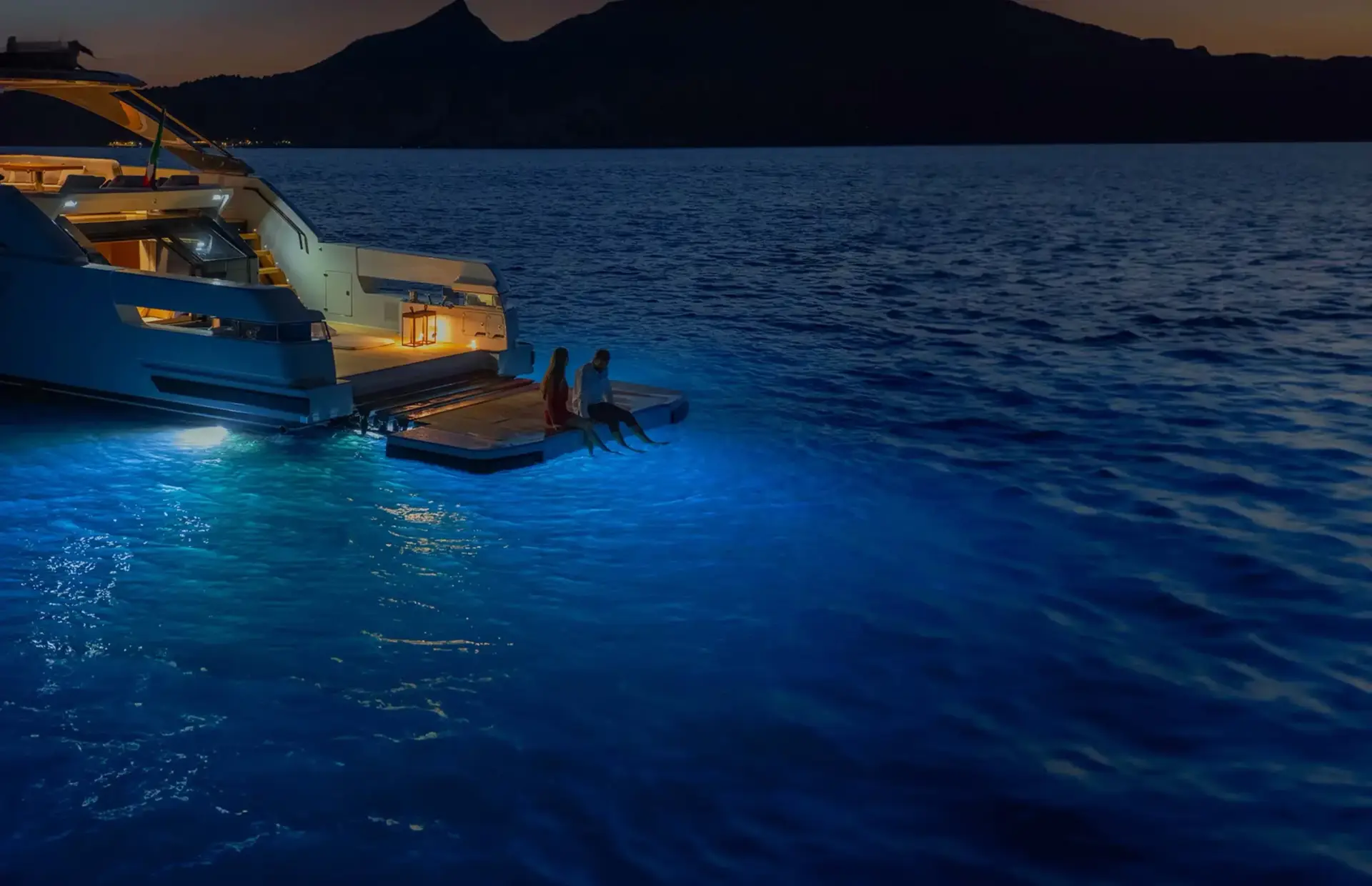 A couple on a yacht at night, with blue lights and mountains.