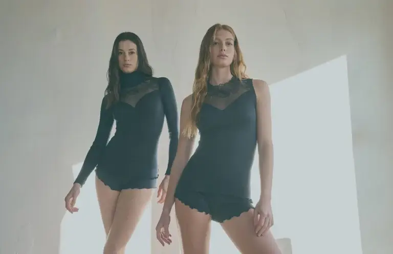 Two women in black lace tops and shorts pose by a sunlit wall.