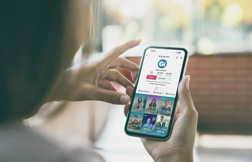 Persona che tiene in mano il telefono e mostra il profilo TikTok e le miniature dei video.