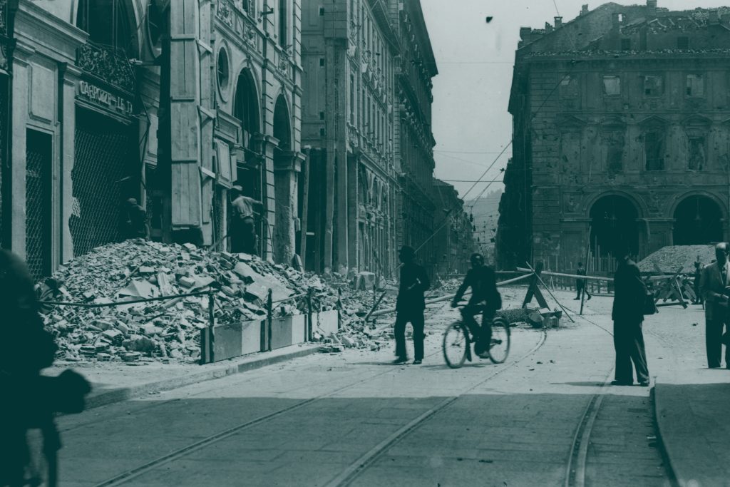 Le persone passano davanti alle macerie di una strada danneggiata fiancheggiata da vecchi edifici.
