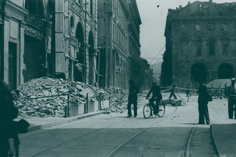 Le persone passano davanti alle macerie di una strada danneggiata fiancheggiata da vecchi edifici.