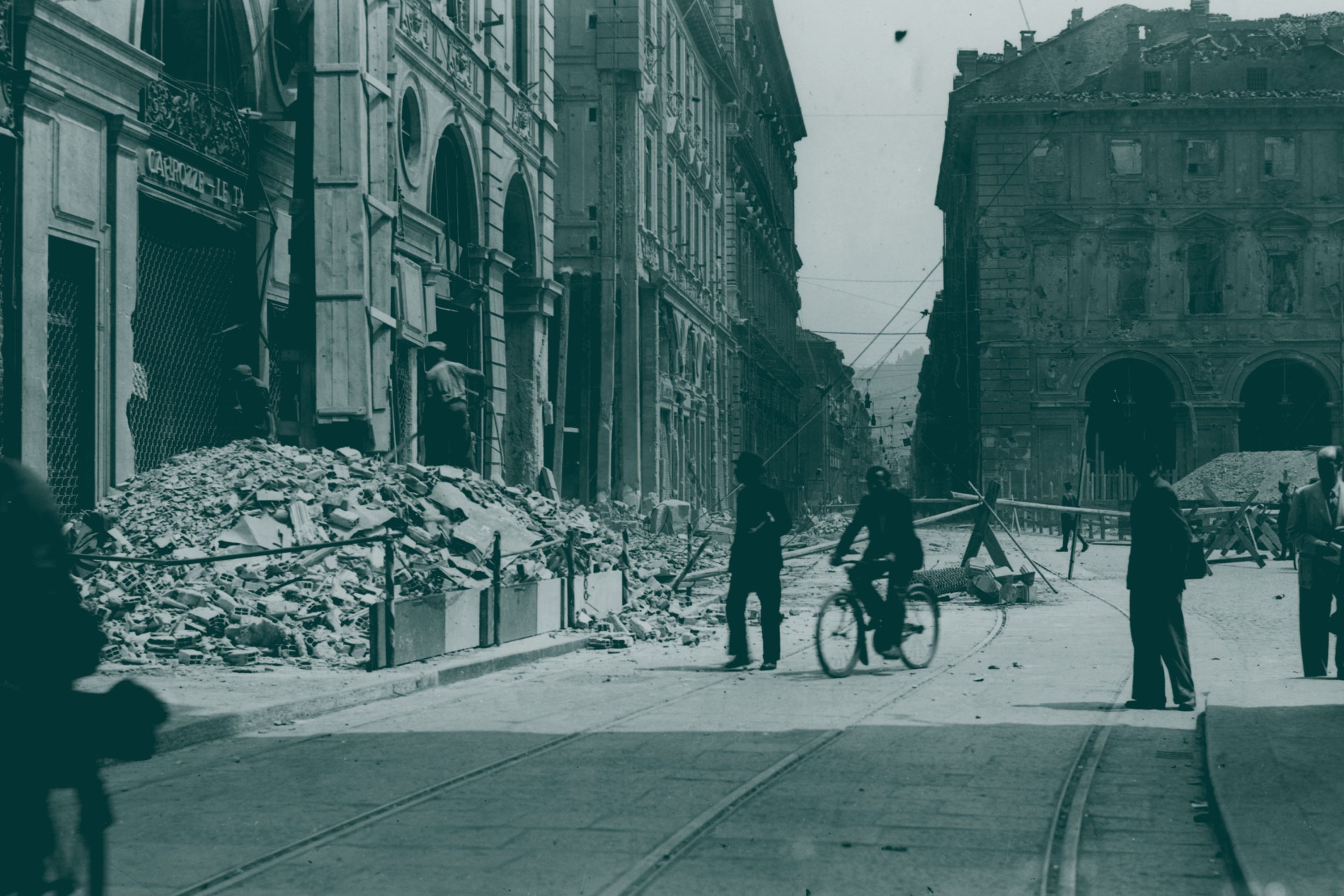 People walk, bike on a city street lined with damaged buildings.