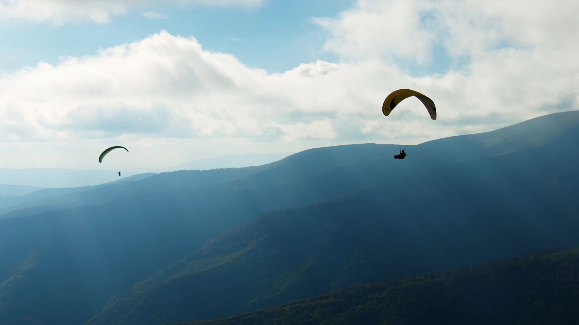 Paragliders soar over sunlit mountains beneath cloudy skies.