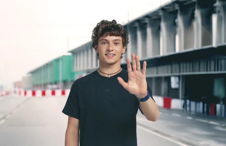 Curly-haired man waves at camera outside by building barriers.